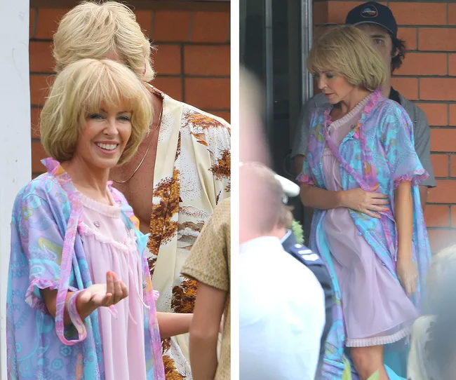 Kylie Minogue dressed in pastel robe and nightgown on a movie set, smiling and gesturing with her hand.