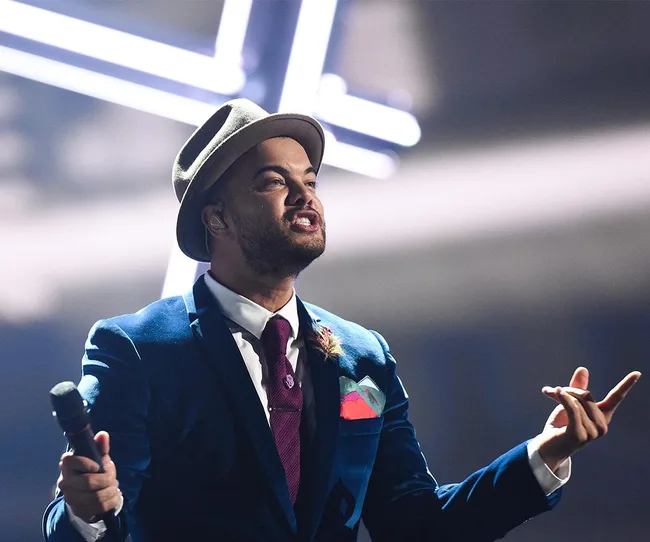 A person in a suit and hat passionately singing on stage with a microphone.