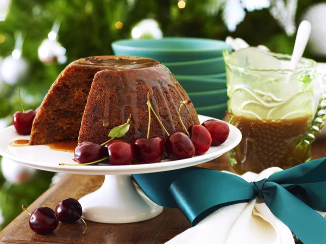 Christmas pudding with cherries on a white stand, green plates, and brown sauce in a glass pitcher, festive background.
