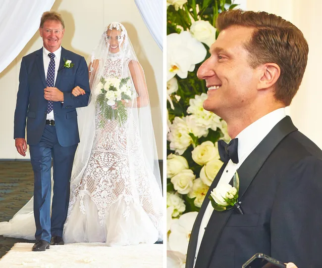 Bride in ornate lace dress walking with a man; groom smiles. White floral decor surrounds them in a wedding ceremony.