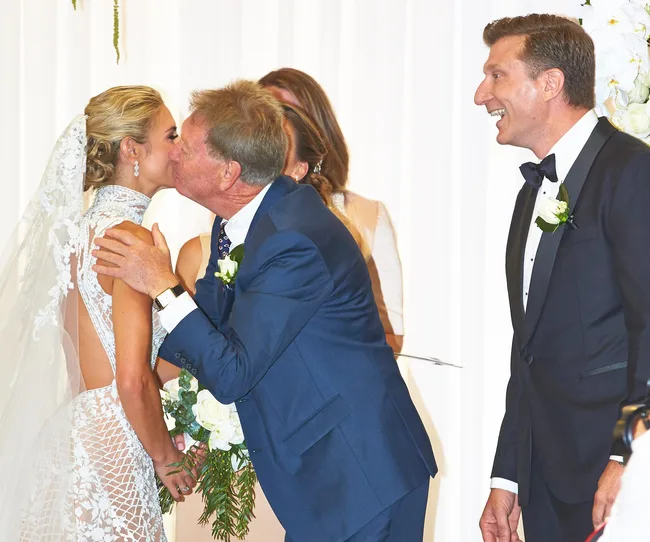 Bride in lace gown kisses older man in blue suit, with groom in tuxedo smiling nearby at wedding ceremony.