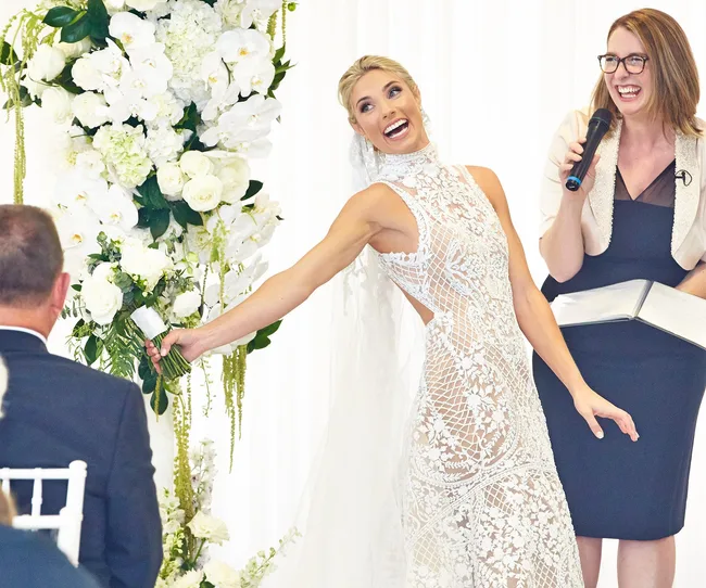 Bride in lace dress holding bouquet, smiling at officiant during wedding ceremony, surrounded by white floral arrangements.
