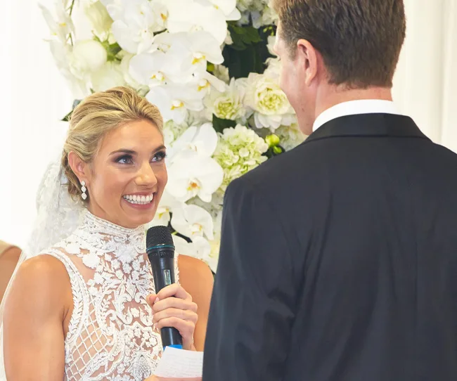A bride in a lace dress holds a microphone and smiles at a groom during a wedding ceremony with floral decorations.