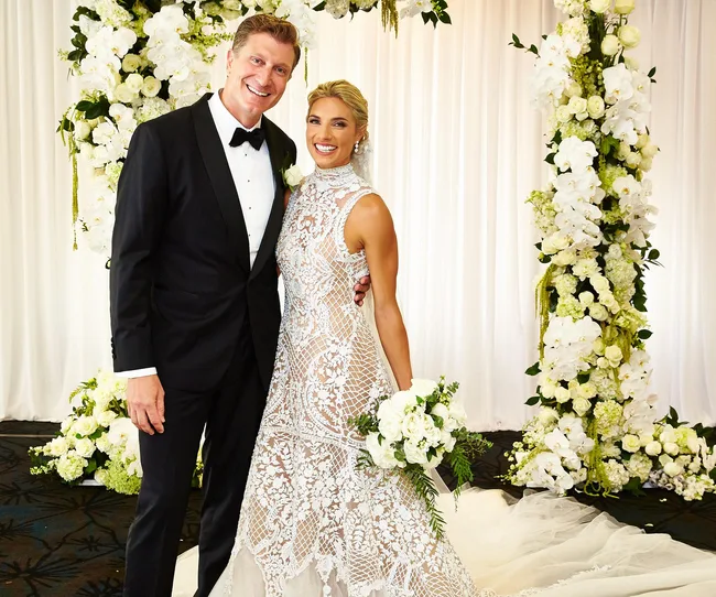 Couple in wedding attire posing smilingly in front of floral arch. The bride holds a white bouquet.