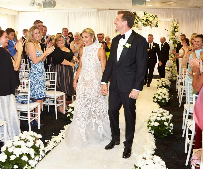 A bride in a lace gown and a groom in a tuxedo walking down the aisle, guests applauding joyfully.