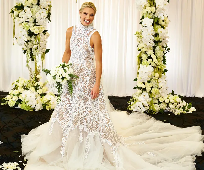 Bride in an intricate lace wedding gown holding a bouquet, surrounded by white floral arrangements.