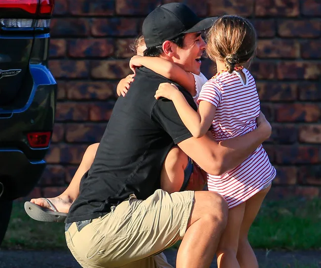 Man in black cap hugging two children near a brick wall, one child wearing a striped outfit.