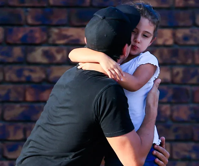 A man in a black shirt and cap hugs a young girl against a brick wall background.