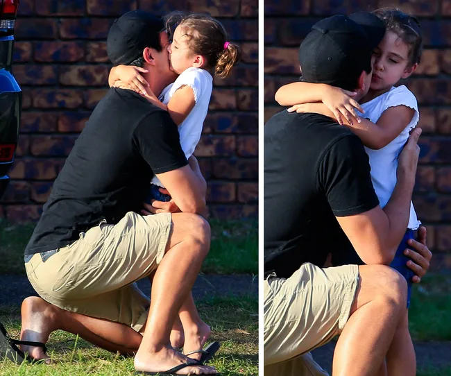 A man in a black cap and shirt kneels to hug and kiss a young girl wearing a white shirt in an outdoor setting.