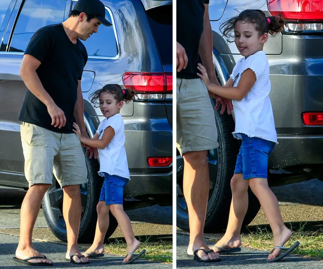Man in black shirt and beige shorts with young girl by a gray SUV, both in flip-flops.