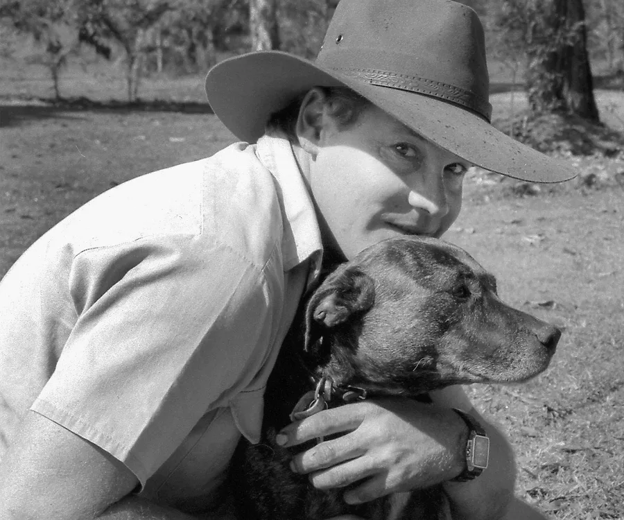 A person in a hat lovingly hugs a dog outdoors in a black and white photo.