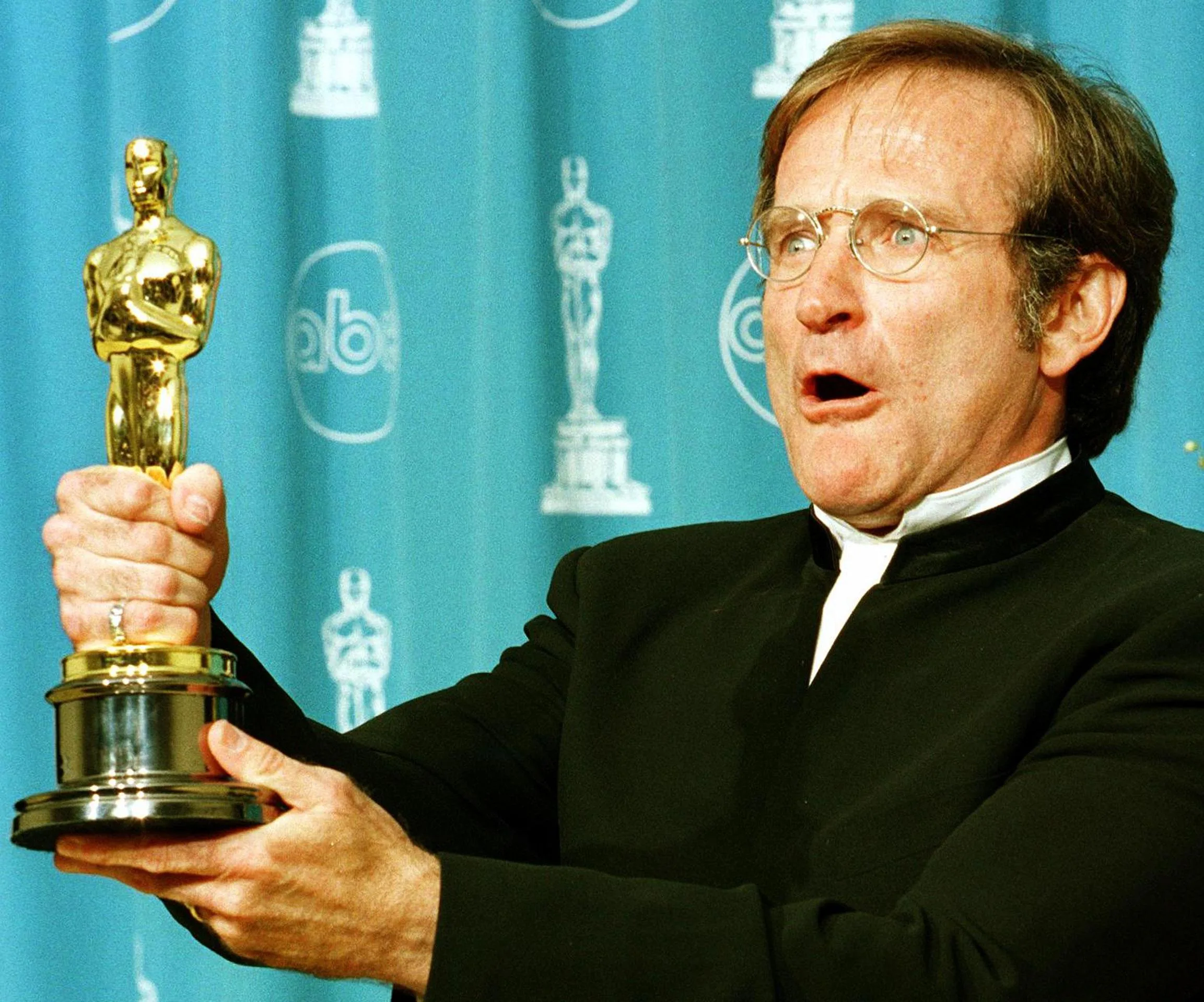 Robin Williams holds an Oscar award with an excited expression at an award ceremony.
