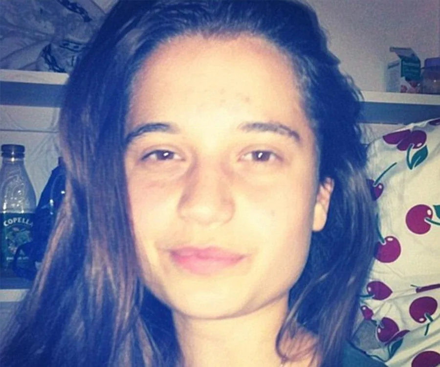Teenage girl with long dark hair smiling softly, indoors with shelves and a pillow in the background.