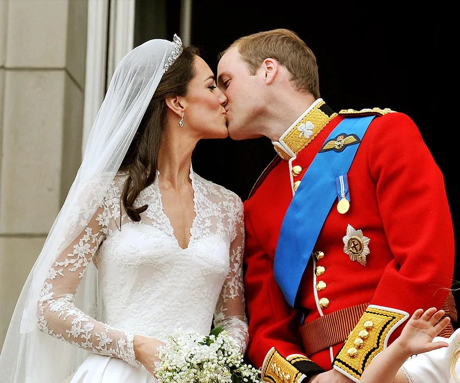 A bride and groom share a kiss on a balcony, with the groom in military attire and the bride in a lace wedding dress.