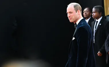 Man in a dark suit with a bow tie smiles, standing in front of a dark background with two people in the distance behind him.