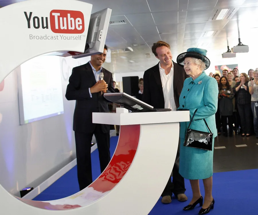 Queen Elizabeth visits a YouTube office, interacting with staff and a podium display screen.