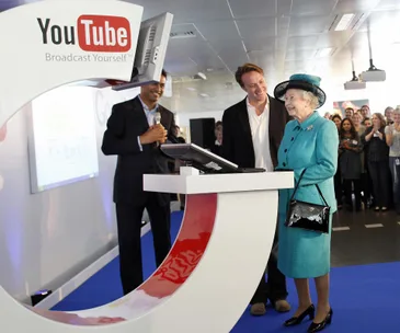 Queen Elizabeth visits a YouTube office, interacting with staff and a podium display screen.