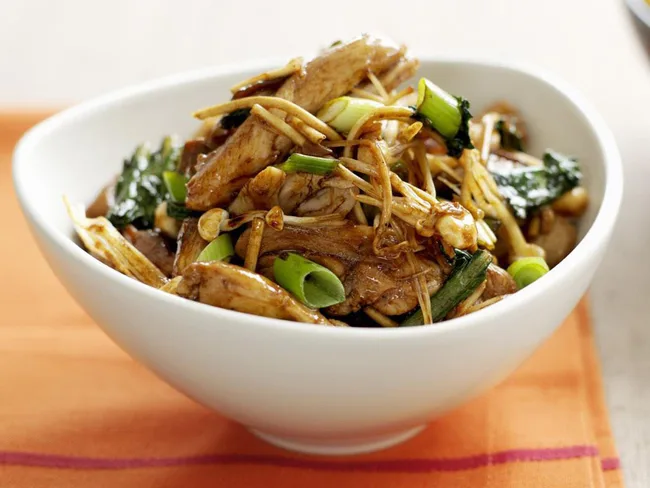 A bowl of cooked stir-fry with chicken, green onions, and vegetables, placed on an orange napkin.