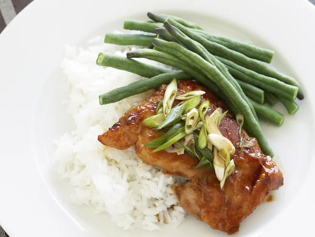 Grilled chicken with glaze, topped with scallions, served over white rice and a side of green beans on a white plate.