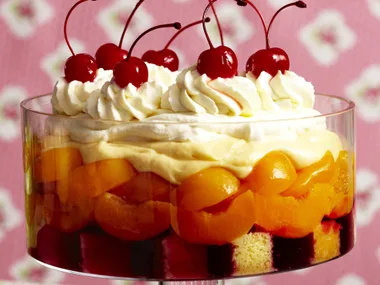 Layered trifle dessert in glass dish with fruit, cream, and cherries on top, against a floral pink background.