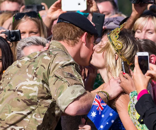 A man in military uniform kisses a woman in a crown, holding an Australian flag, surrounded by people taking photos.