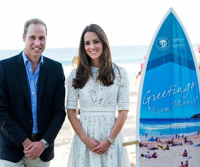 Two people smiling next to a surfboard that says "Greetings from Manly" on a beach.