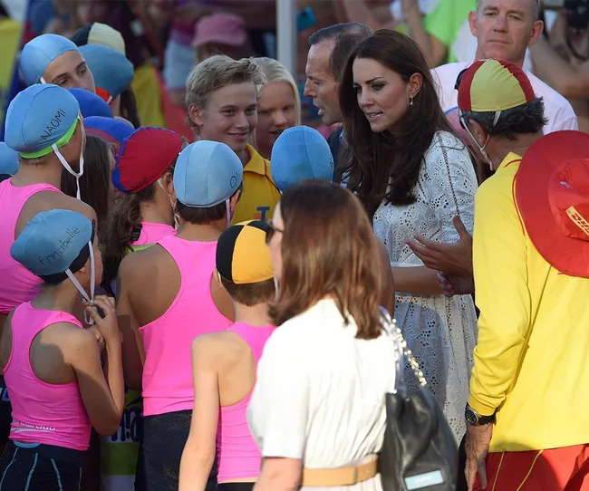 A woman in a white dress interacts with a group of kids in pink swim caps and shirts at an outdoor event.