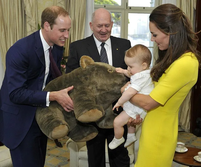 Man shows a large stuffed animal to a woman holding a baby while an older man looks on, in a formal room.