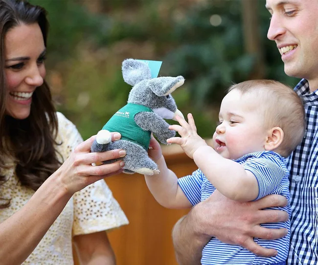 A smiling woman holds a plush toy as a baby reaches for it, held by a smiling man outdoors.