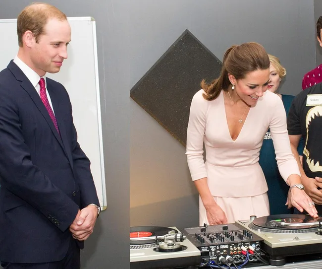 A woman learns to DJ on a turntable as a man in a suit watches nearby.
