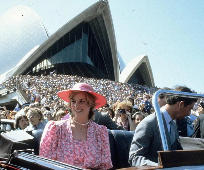A woman in a pink outfit and hat smiles in a car near the Sydney Opera House with a large crowd in the background.