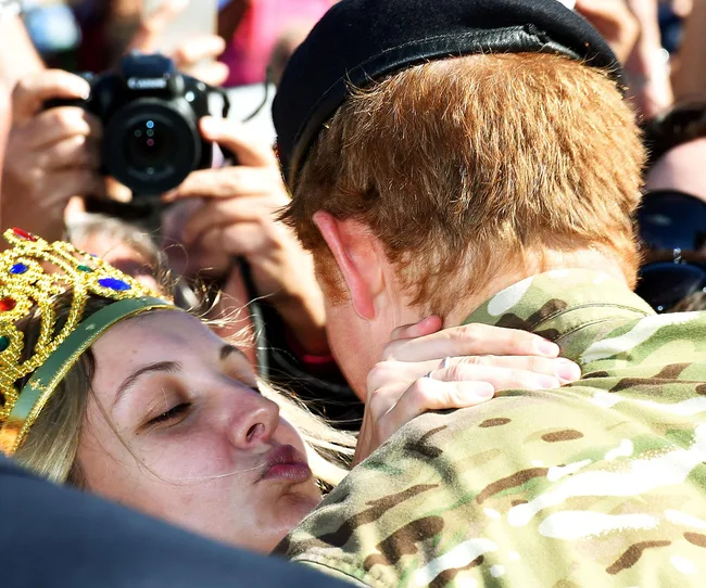A person in military uniform receives a kiss from a woman wearing a crown, with cameras capturing the moment.