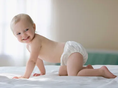 A smiling baby in a diaper is crawling on a bed in a softly lit room.