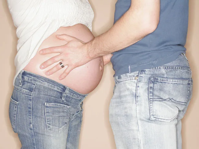 Pregnant person in jeans, wearing a white shirt, with partner's hands on belly, both in casual denim attire.