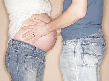 Pregnant person in jeans, wearing a white shirt, with partner's hands on belly, both in casual denim attire.