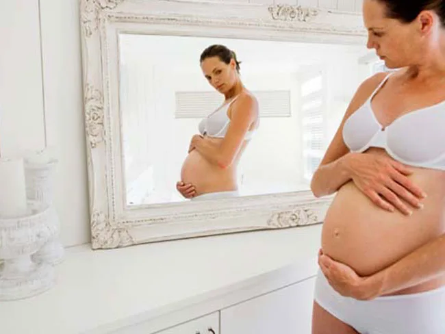 Pregnant woman in underwear at 33 weeks, holding stomach and looking at reflection in a white framed mirror.