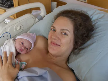 A woman in a hospital bed holds a newborn baby, wrapped in a white blanket, wearing a pink hat, close to her chest.