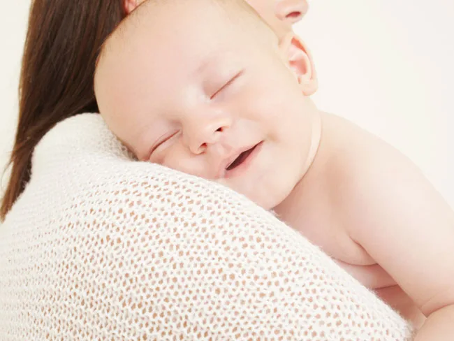 Smiling baby rests on a woman's shoulder, wrapped in a knitted blanket.