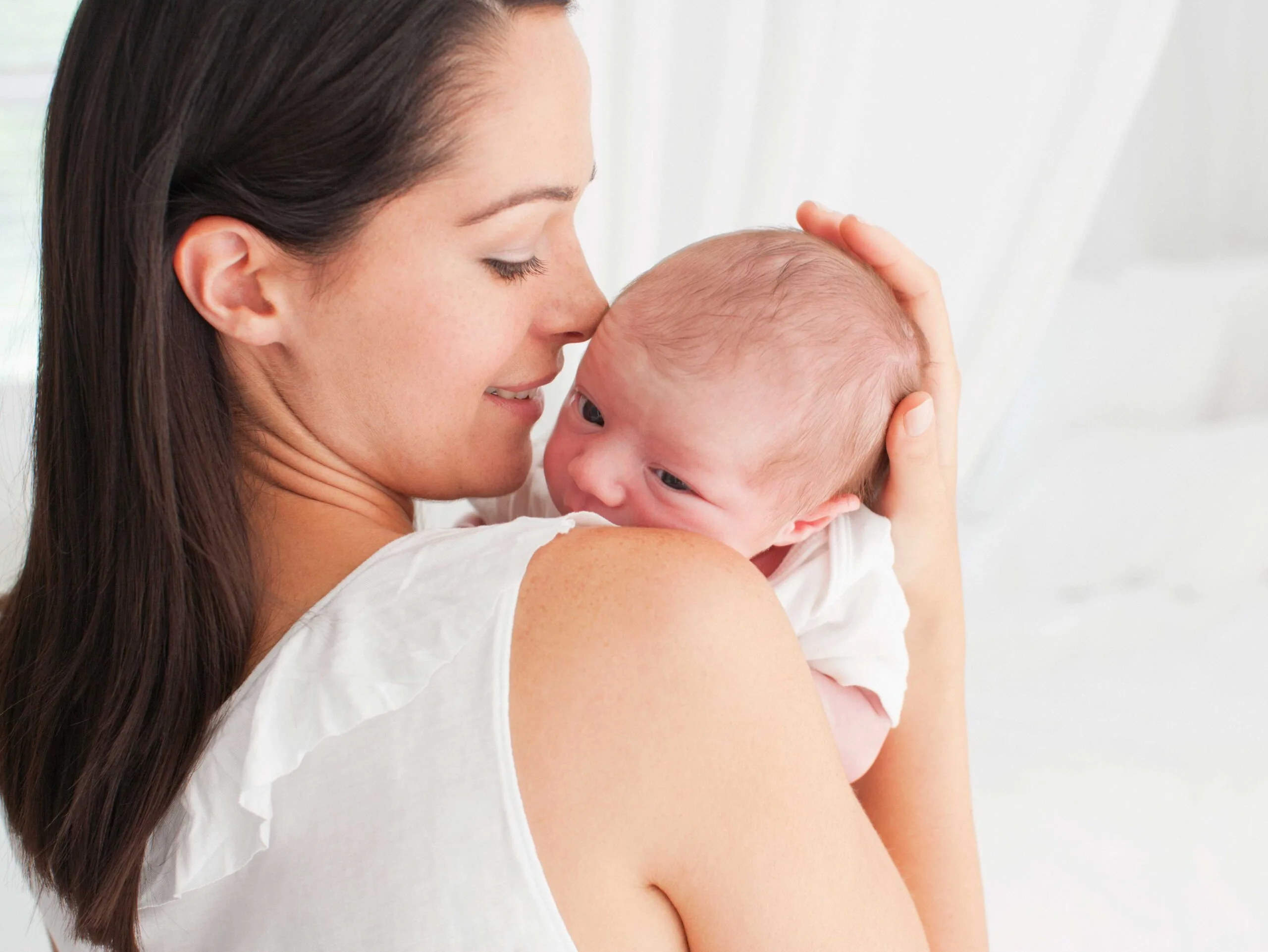 Mother cuddling newborn close to her shoulder, both looking peaceful in a bright, serene setting.