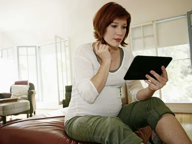 Pregnant woman sitting on a couch, reading from a tablet in a well-lit living room.