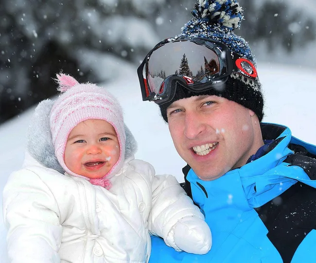 Father in blue snow jacket holds smiling toddler in pink hat, snowy background.