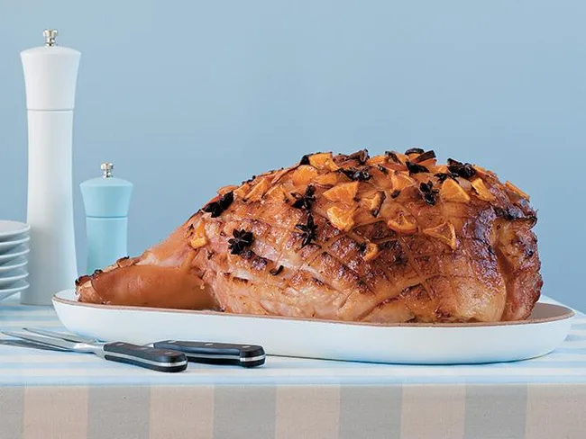 Glazed ham studded with cloves and orange slices on a white platter, with cutlery and salt shaker beside.