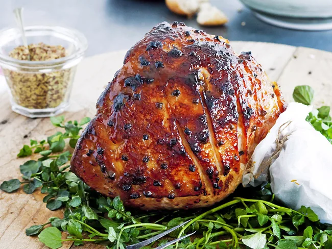 Roast ham glazed with dark caramelized sauce, garnished with fresh greens on a wooden board, jar of mustard nearby.
