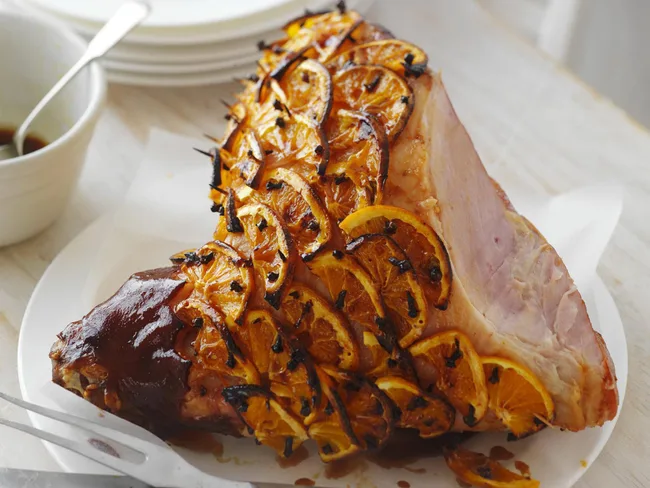 Glazed ham decorated with orange slices and cloves on a white plate.