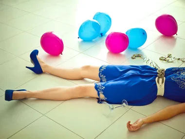 Person in blue dress and heels lying on floor surrounded by pink and blue balloons, with overturned glass nearby.