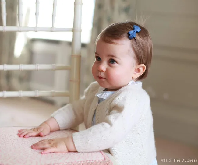 A young child with a blue bow in their hair looks sideways, wearing a white sweater, in a softly lit room.