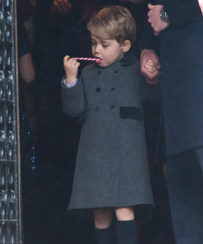 Young child in a grey coat holds a candy cane, standing at a Christmas church service, holding an adult's hand.