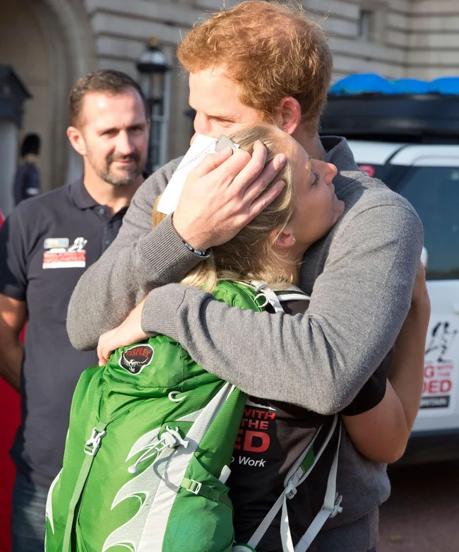 Prince Harry warmly hugs a soldier with a green backpack, against a backdrop of onlookers and a vehicle.