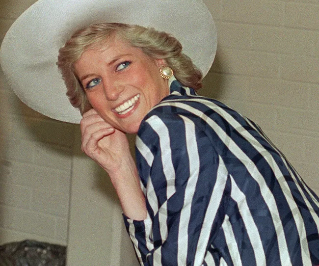 A woman smiling in a wide-brimmed white hat and striped outfit, with short blonde hair and elegant pearl earrings.