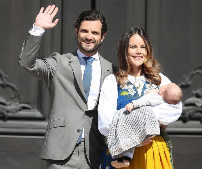 A couple smiling, man waving, woman holding a baby wrapped in a gray blanket, standing outside a building.